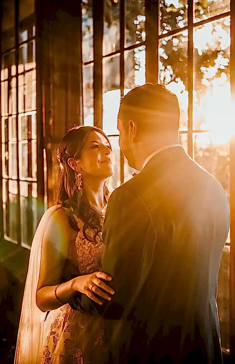 Foto de matrimonio parado junto a ventana por la que entran los rayos de sol